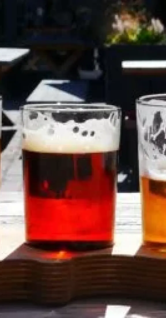 Five beer tasting glasses with different amber-colored beers served on a wooden flight paddle outdoors.